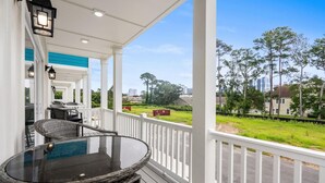 A covered porch with wicker furniture and a glass table, overlooking a green landscape and buildings. - Spacious 4 Bedroom 3.5 Bathroom walking distance to the beach. Close proximity! (Myrtle Beach)