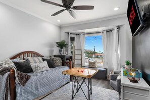 Ein Schlafzimmer mit Bett, Schreibtisch, Deckenventilator, Fernseher und einem Fenster mit Vorhängen.