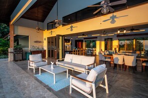 Un espace de vie extérieur comprenant un canapé, des fauteuils, une table à manger, des ventilateurs de plafond et un barbecue.