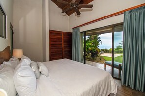 Une chambre dotée d’un grand lit, d’une tête de lit en bois, d’un ventilateur de plafond et offrant une vue sur un jardin grâce à une porte-fenêtre coulissante.