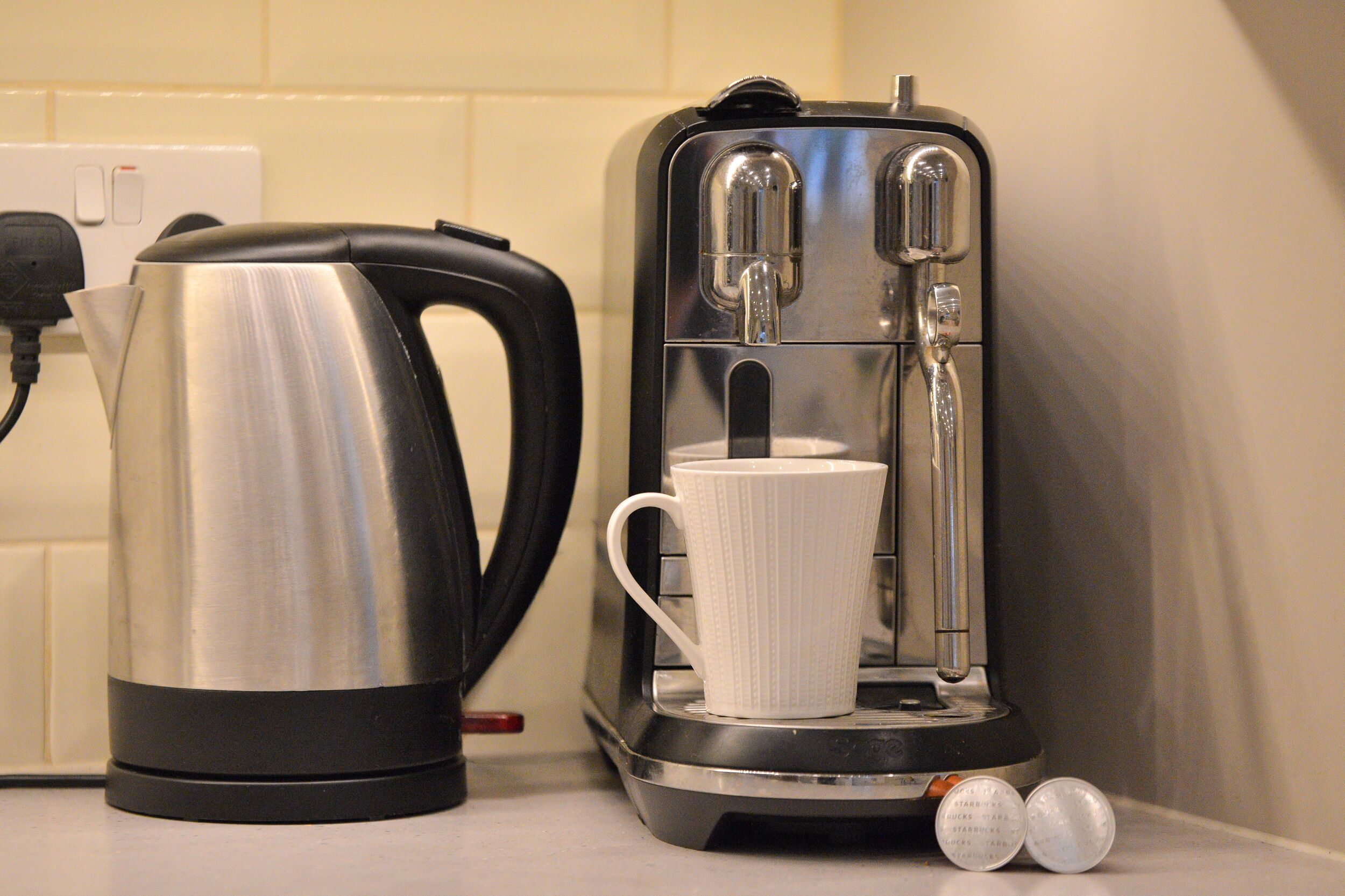 A coffee maker with a white mug on its drip tray, a kettle, and a power outlet in the background.