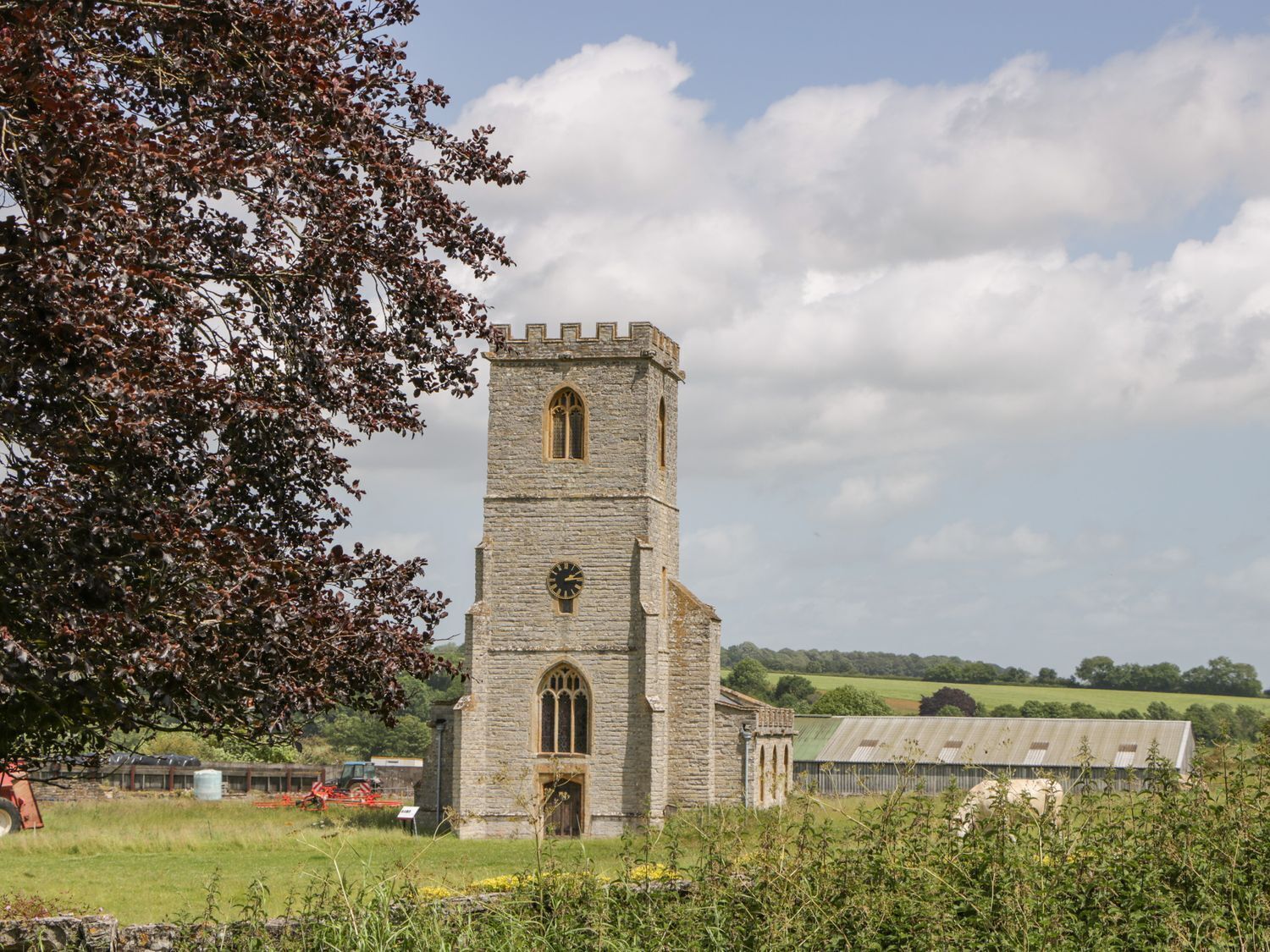A stone church with a clock tower, surrounded by fields and a tree.