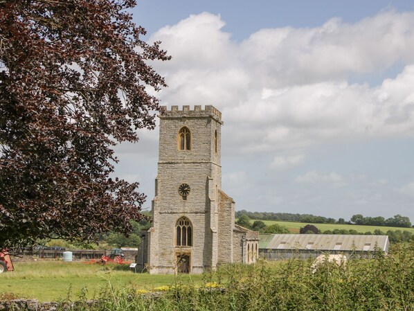 A stone church with a clock tower, surrounded by fields and a tree.
