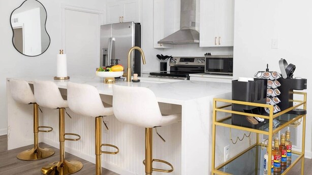 Une cuisine moderne avec des meubles blancs, un réfrigérateur en acier inoxydable, une cuisinière à gaz et un four à micro-ondes. On y trouve trois tabourets de bar blancs aux pieds dorés, un îlot central blanc doté d’un évier, et un chariot de bar noir et doré avec des bouteilles.