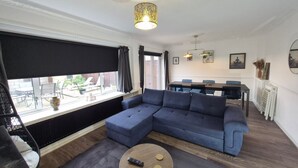 A living room with a sectional sofa, a glass coffee table, a dining area with a table and chairs, and a large window with blinds.