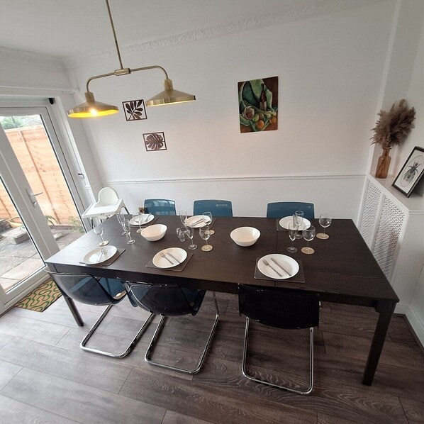 A dining area with a dark wooden table set for six, surrounded by black chairs with chrome legs. There are two wall-mounted pendant lights above the table.