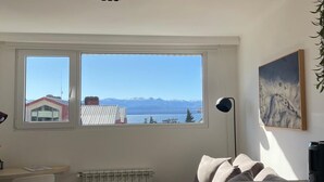 Una habitación con un amplio ventanal que ofrece vistas a montañas y a una masa de agua. Se observa un radiador, un escritorio y una silla.