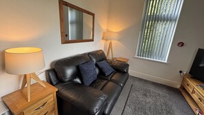 A living room with a black leather sofa, two wooden nightstands with lamps, a large mirror, and a window with blinds.
