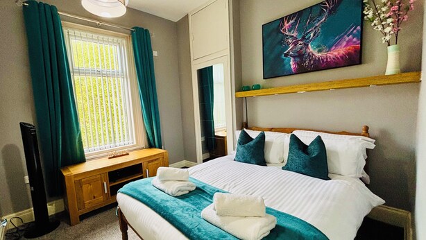 A neatly made bed with a teal blanket and pillows, a wooden bedside table with a lamp, a window with curtains, and a painting on the wall.