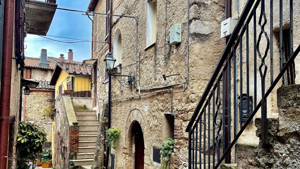 Uma rua de paralelepípedos estreita, ladeada por edifícios históricos de pedra, com portas em arco e uma escadaria que leva ao segundo andar.