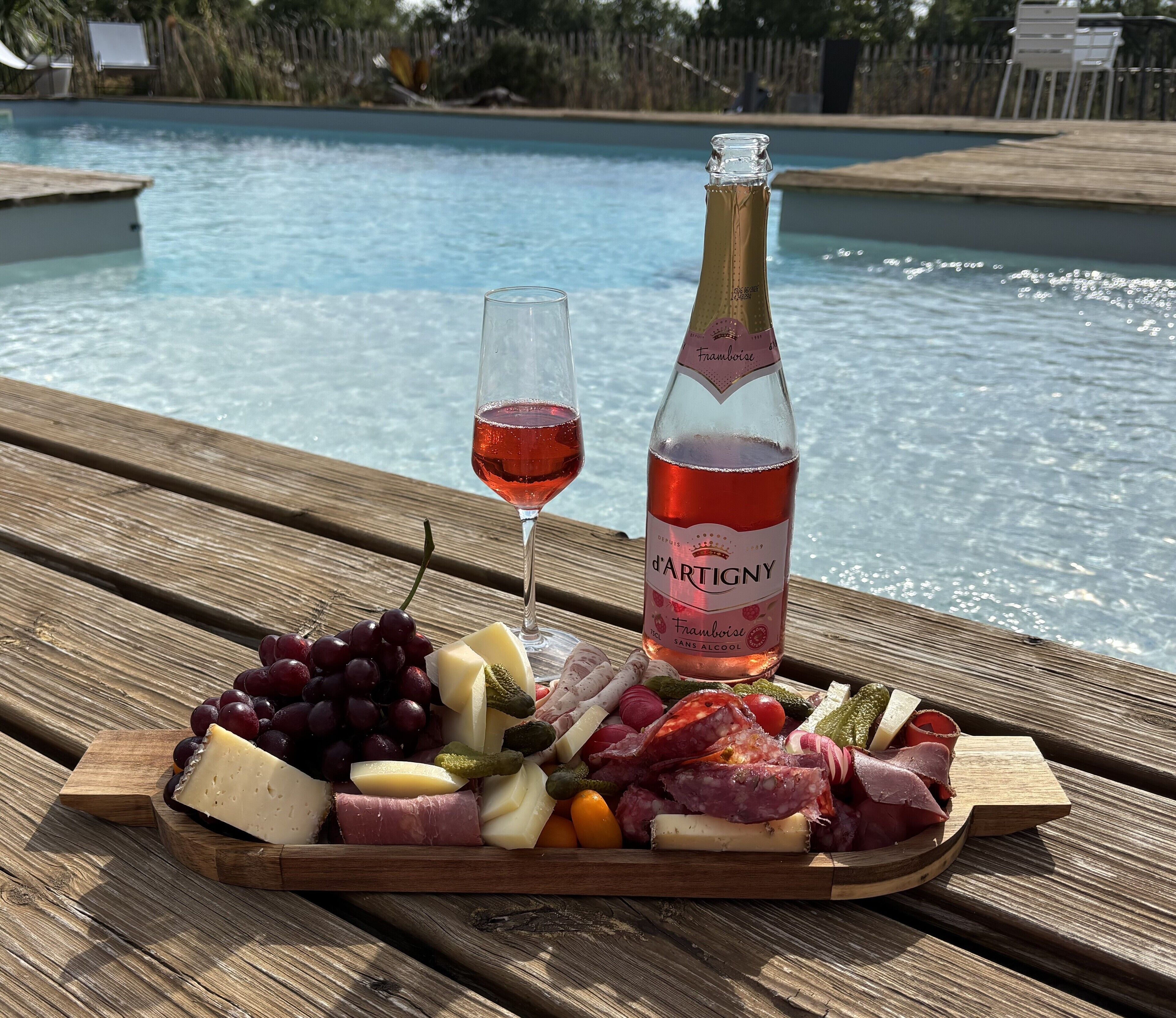 Un montaje para picnic con una botella de vino d'Artigny, una copa de vino tinto y una tabla de fiambres junto a una piscina.