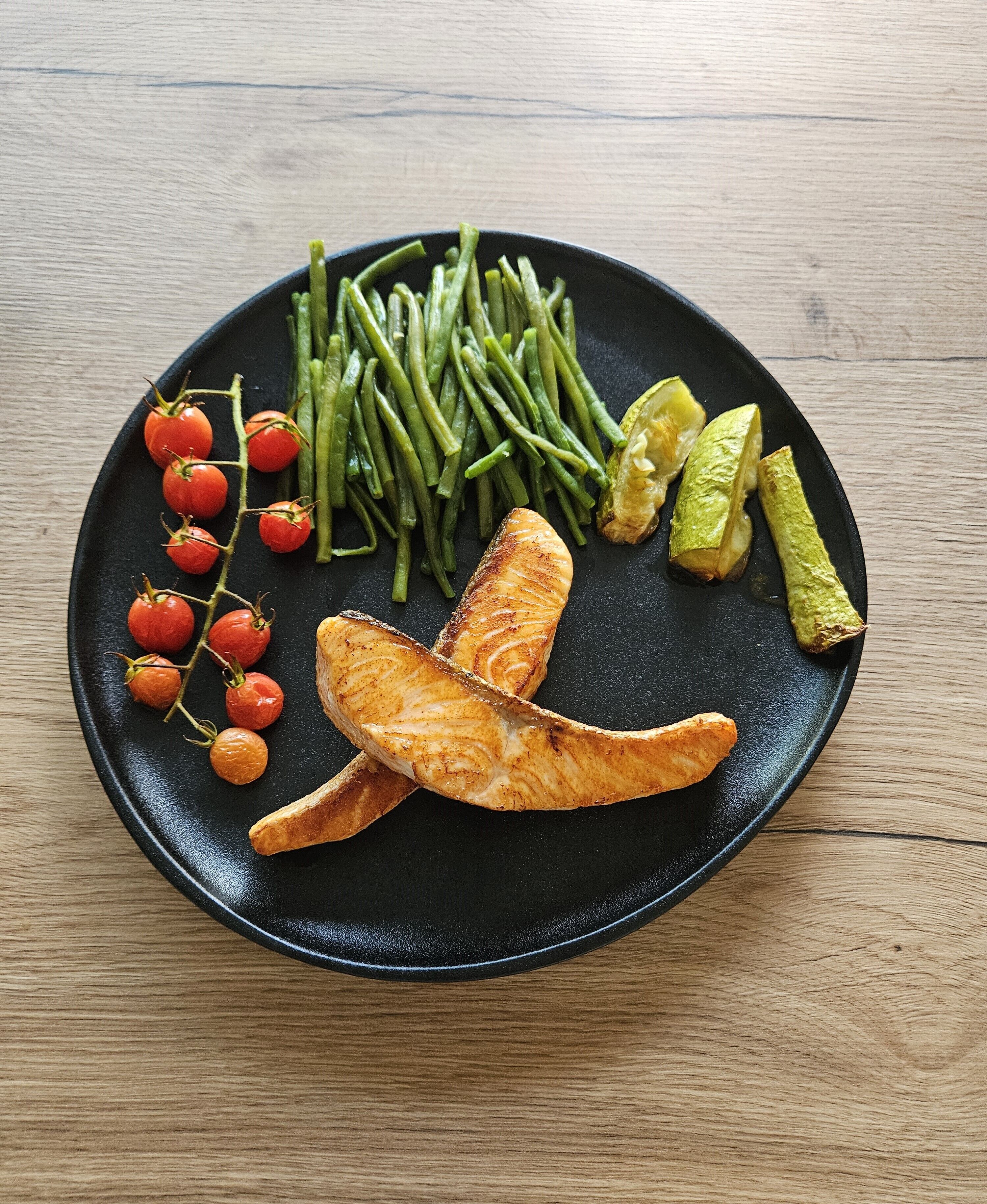 Une assiette noire contenant du saumon grillé, des haricots verts, des tomates cerises et des concombres marinés, le tout posé sur une table en bois.