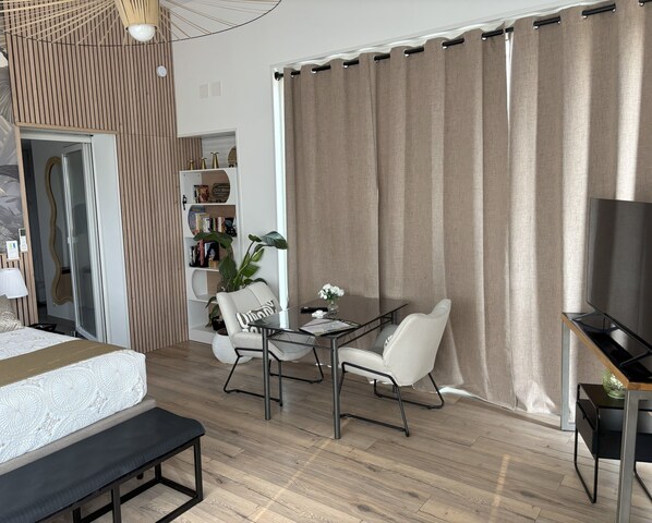 A modern hotel room with a wooden floor, a bed with a white headboard, a desk with a TV, a bookshelf, and a large window with curtains.