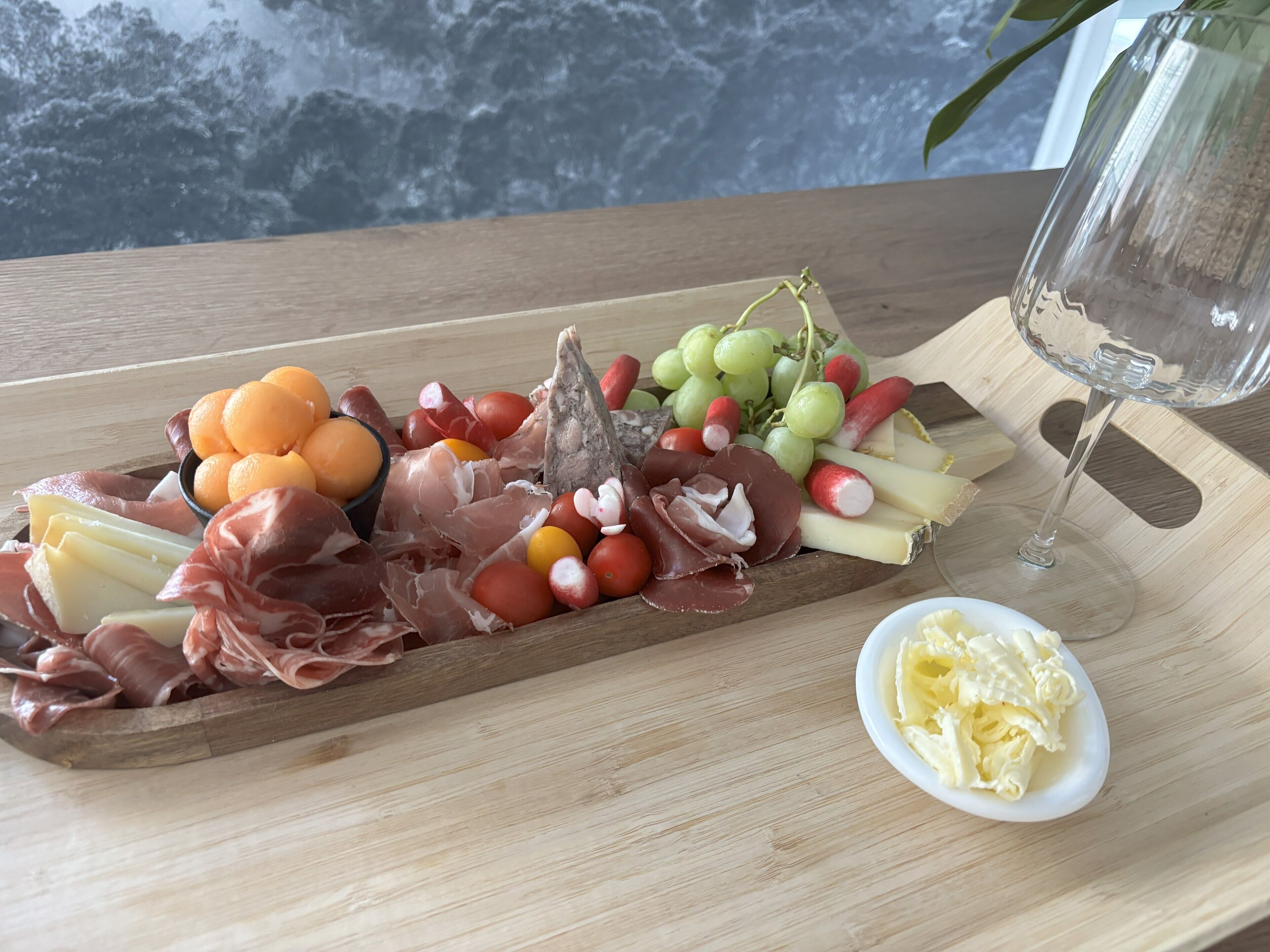 Una tabla de madera con varios quesos, embutidos, frutas y una copa de vino.