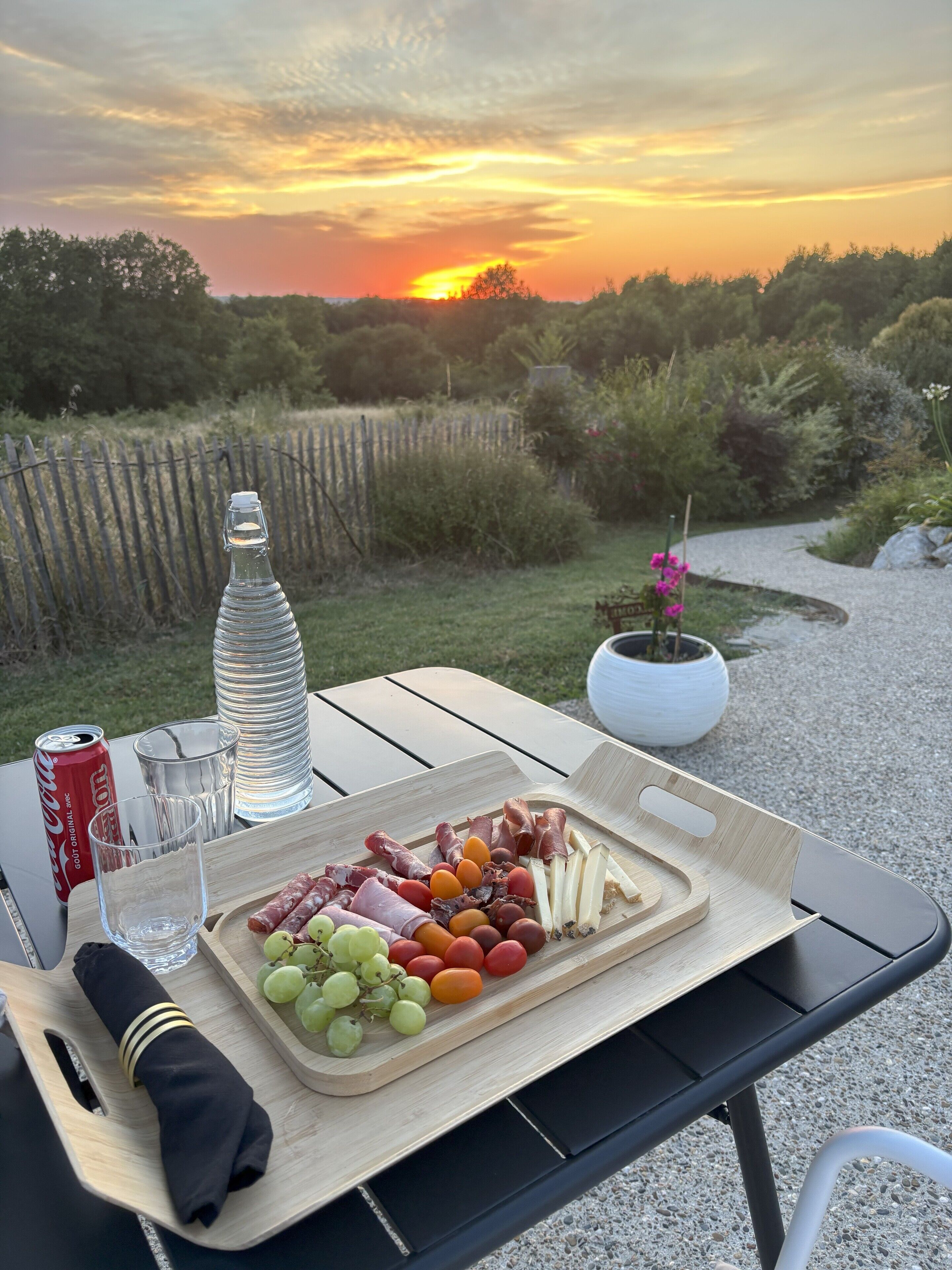 Een picknickopstelling met een houten tafel, een fles, een blikje en een schaal met eten, waaronder druiven, tomaten en vlees.