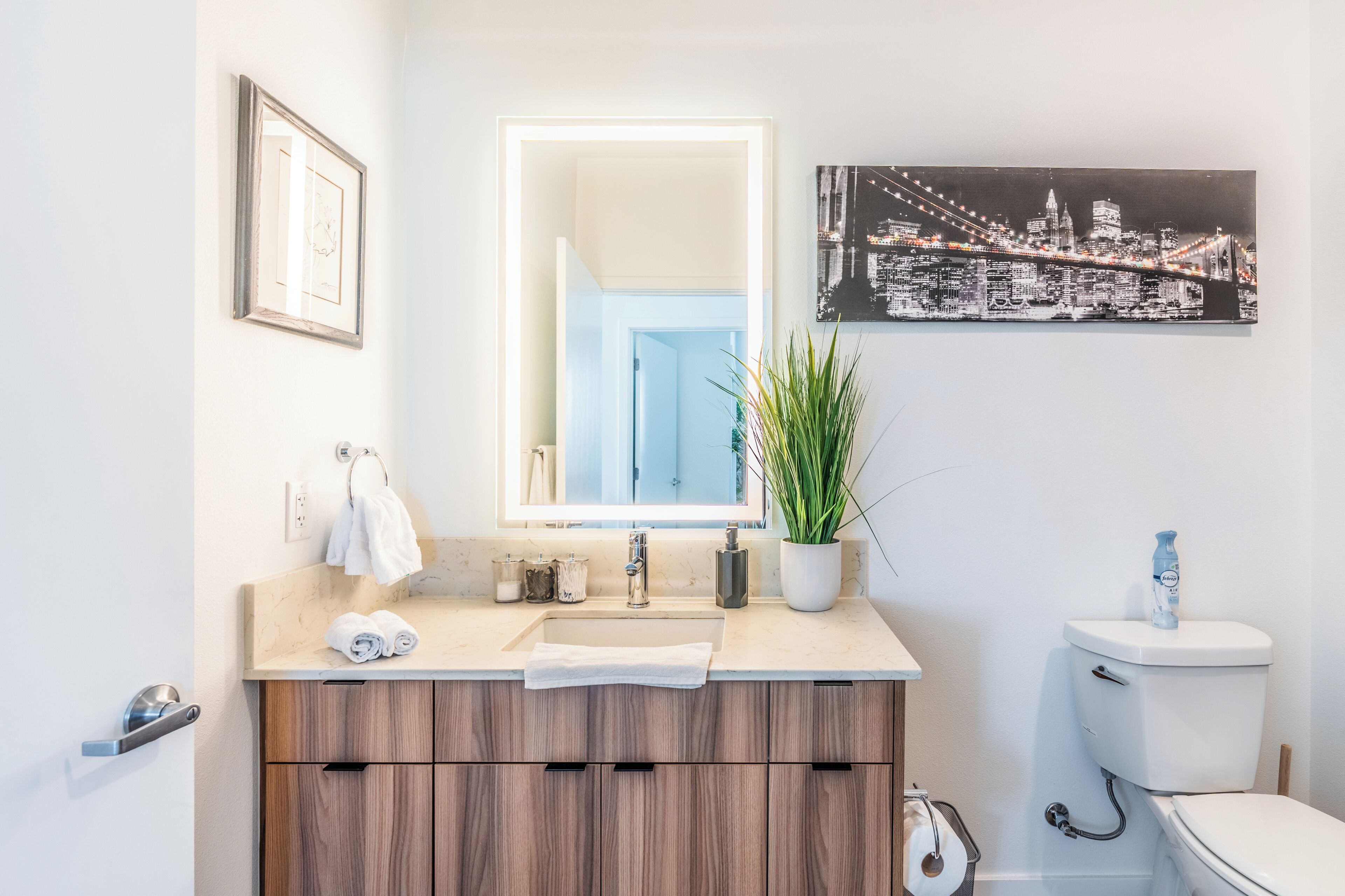A bathroom with a sink, mirror, toilet, and a framed cityscape picture.
