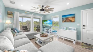 A spacious living room with a large sofa, a coffee table, and a dining area with a white table and chairs. There is a ceiling fan, a TV, and a painting on the wall.
