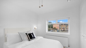 Une chambre avec un grand lit, des murs blancs, une fenêtre donnant sur des immeubles et un radiateur.