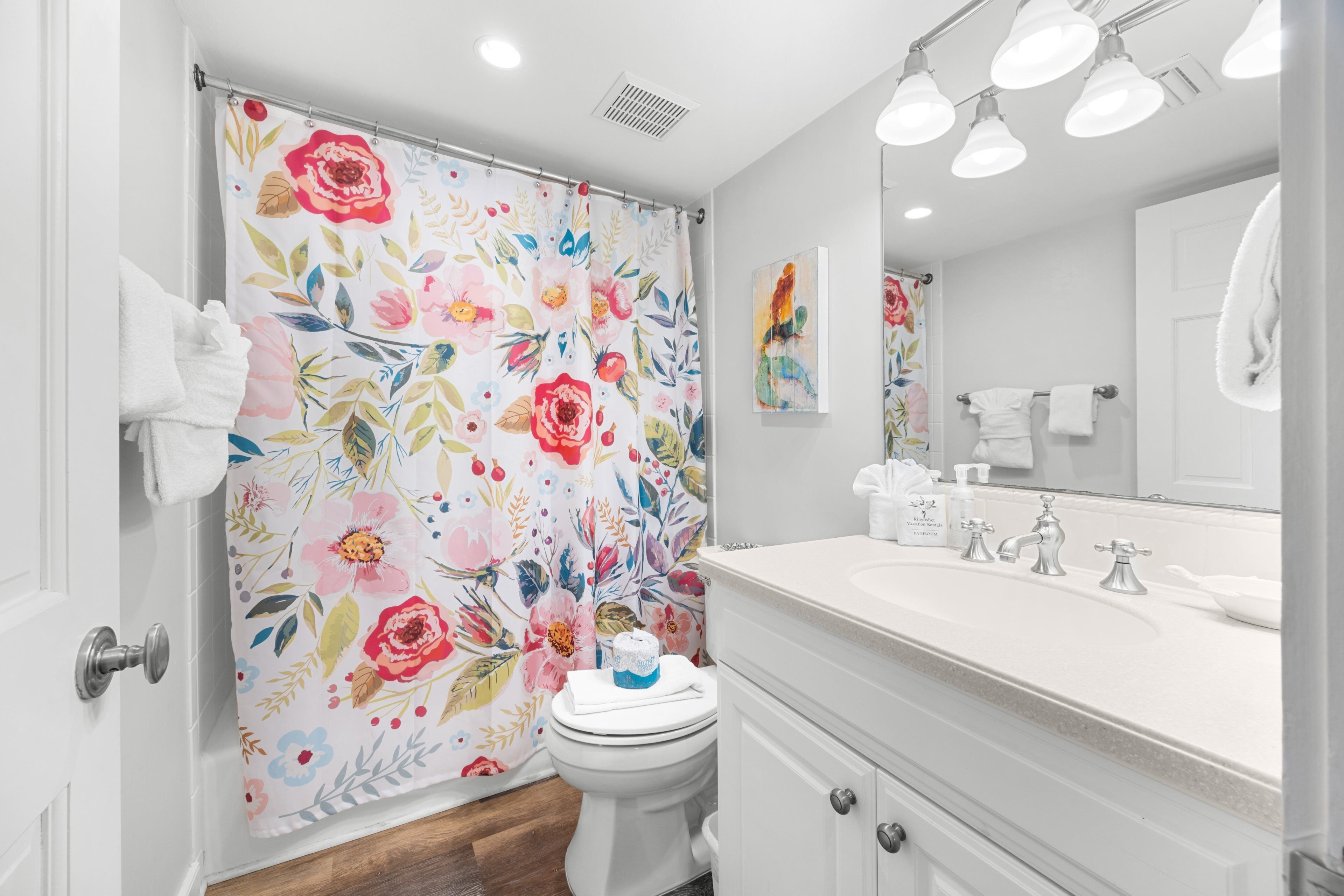 Ein Badezimmer mit blumigem Duschvorhang, Toilette, Waschbecken mit Spiegel und Handtuchhalter.