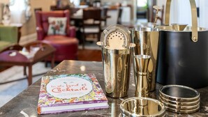 Une table sur laquelle se trouve un livre intitulé « Au pays des cocktails », un shaker à cocktails et un pot noir doté d’anses dorées.