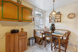 Une cuisine avec des meubles de rangement en bois, une table à manger avec des chaises, une poubelle et une fenêtre avec des rideaux.