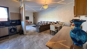 Une cuisine moderne dotée d’un four encastré, un coin repas avec une grande table et des chaises, et un salon avec un canapé et des coussins.