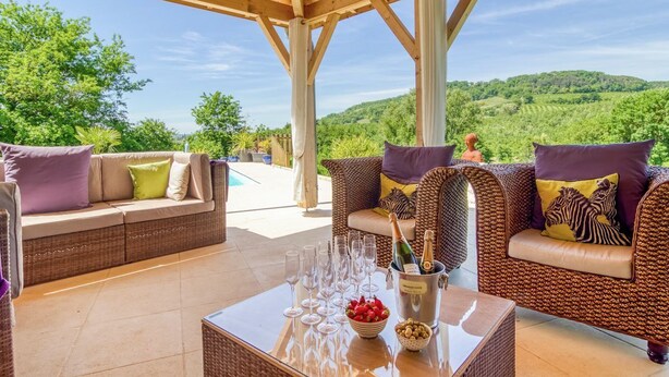 A covered outdoor seating area with wicker furniture, a glass table with wine glasses and a bottle, and a view of a pool and greenery. - Manoir Des Cedres - Eight Bedroom Castle, Sleeps 20 (Castelmoron-sur-Lot)