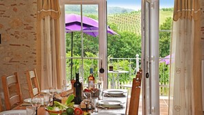 A dining area with a table set for four, a wine bottle, and a view of a vineyard through large windows. - Manoir Des Cedres - Eight Bedroom Castle, Sleeps 20 (Castelmoron-sur-Lot)