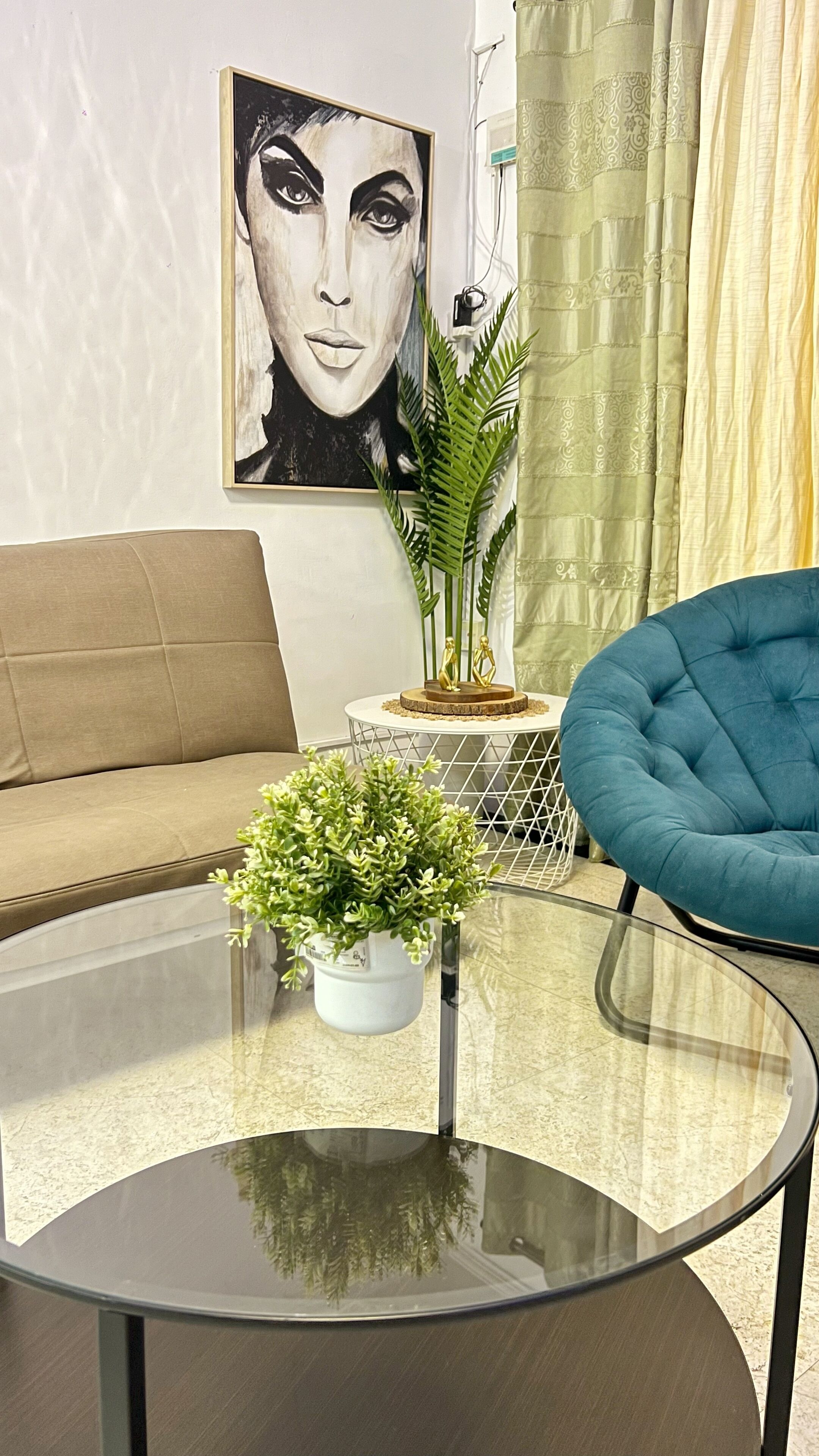 A living room with a sofa, a round glass coffee table with a potted plant, a blue armchair, and a painting on the wall.