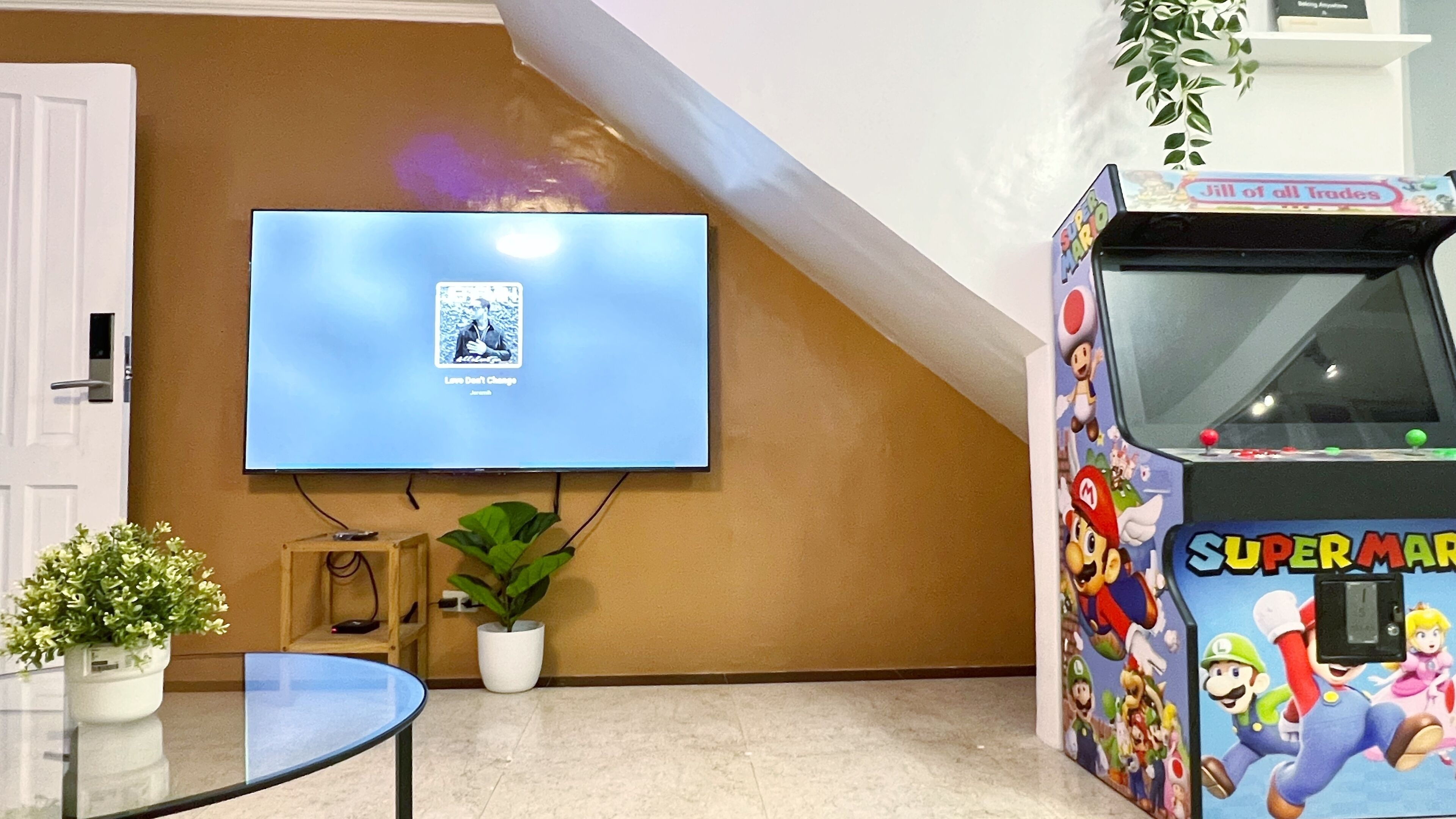 Quarto com televisão presa na parede, um jogo de fliperama com tema do Mario, uma pequena mesa com um vaso de planta e uma porta.