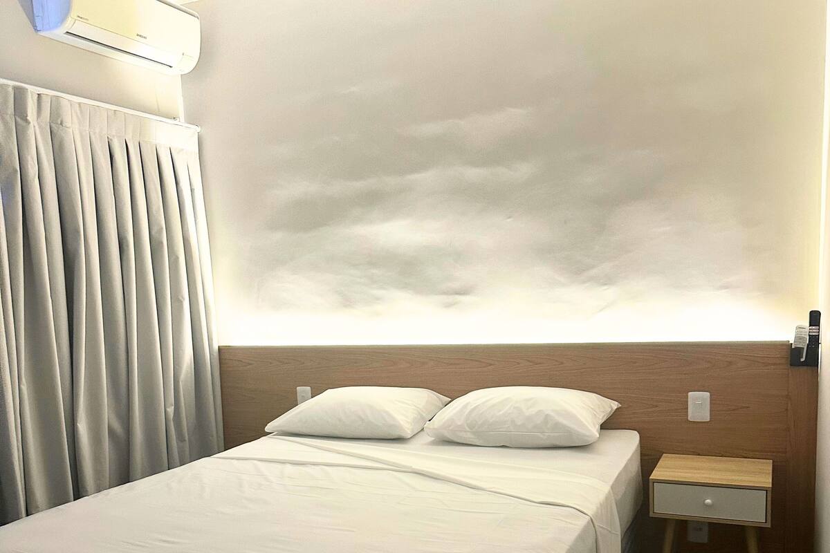 Quarto de hotel com uma cama grande, uma mesinha de cabeceira, ar condicionado na parede e vista do céu pela janela.