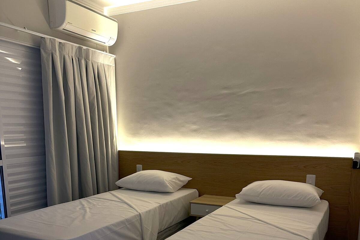 Quarto de hotel com duas camas de solteiro, uma janela grande com cortinas, um aparelho de ar condicionado e uma cabeceira de madeira.