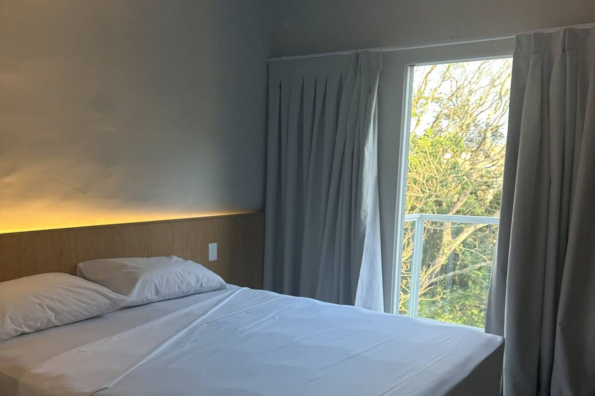 Quarto com cama grande, cabeceira de madeira e vista para árvores pela janela.