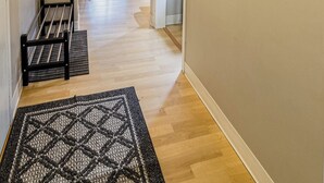 En gang med trægulv, et sort-hvidt tæppe med mønster, en rød taske hængende på en krog, et farverigt abstrakt billede på væggen og udsigt til en balkon med stol og sofa.