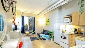 En moderne lejlighed med køkken, spiseområde og opholdsstue. Der er et stort vindue, en sofa, et spisebord og et køkken med komfur og mikroovn.