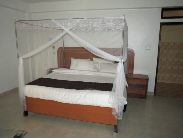 A single bed with a wooden frame and white bedding, a nightstand with a lamp, and a closed door in a plain room. - Kim's Breeze Hotel Ltd and Choma Zone (Narok)