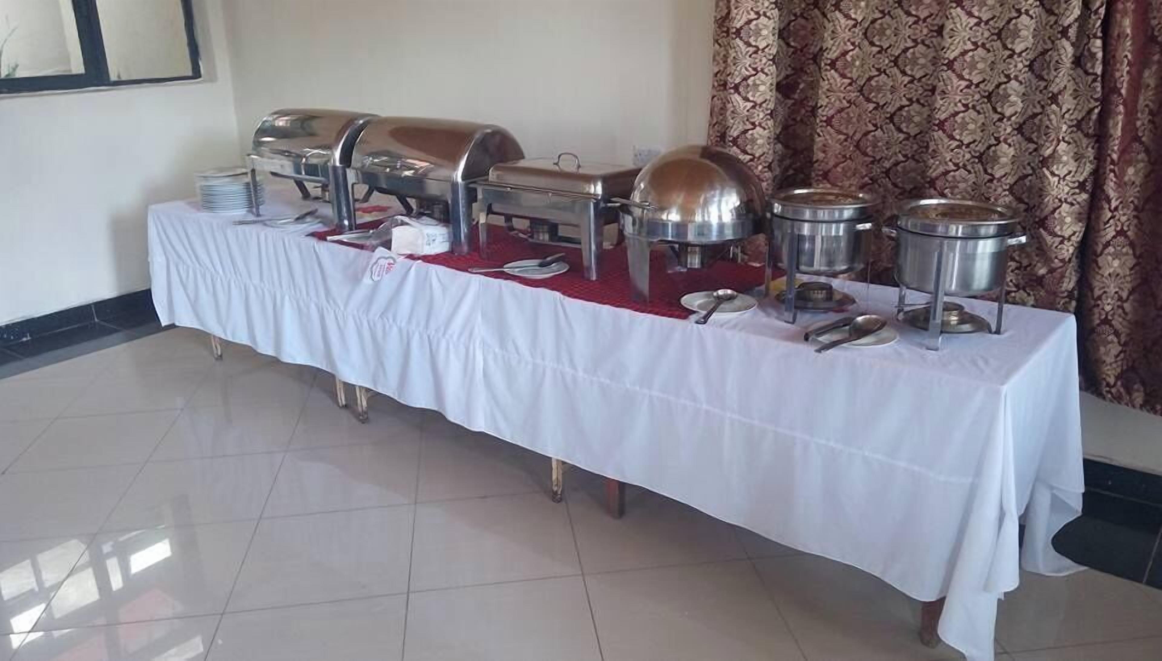 A buffet table with various food containers and utensils, set up in a room with patterned curtains and tiled flooring.
