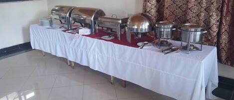 A buffet table with various food containers and utensils, set up in a room with patterned curtains and tiled flooring.