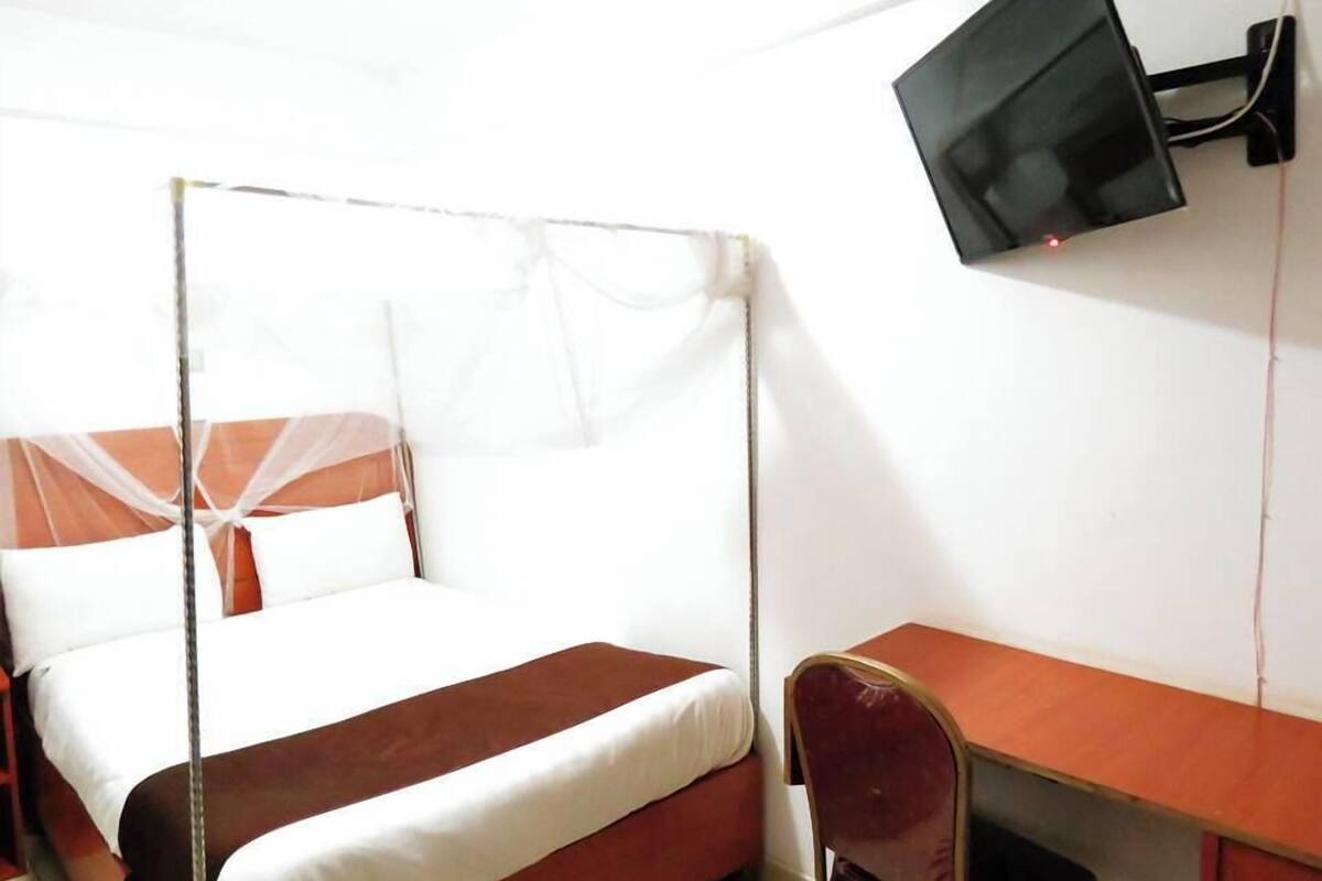Uma cama de solteiro com mosquiteiro, uma escrivaninha e uma televisão fixada na parede.