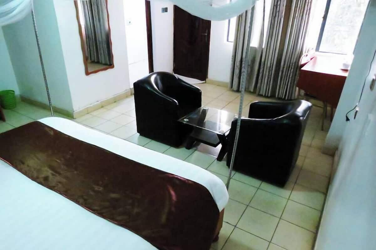 Quarto de hotel com cama, duas cadeiras pretas, uma mesinha e uma janela com cortinas.