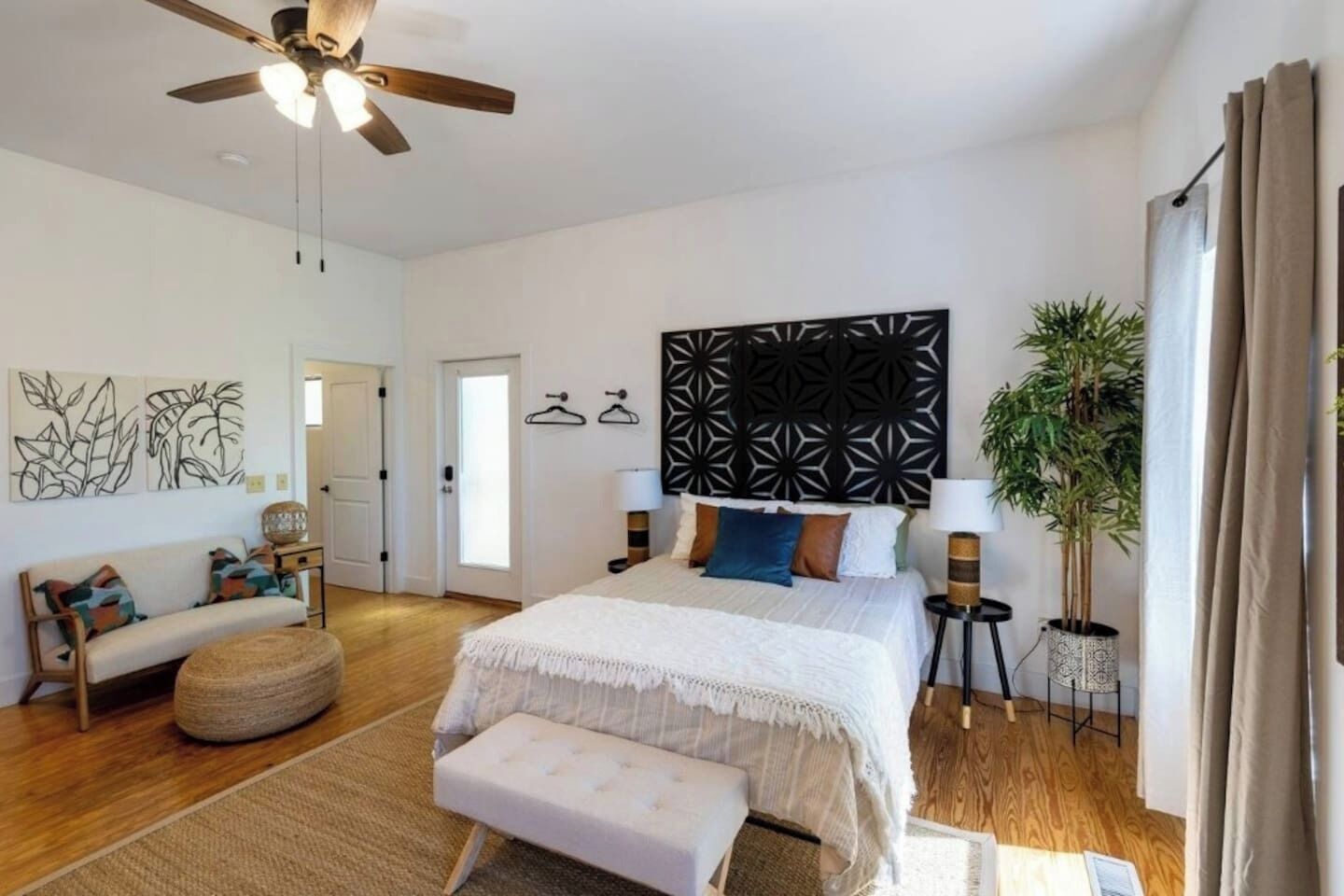 A bedroom with a bed, a ceiling fan, a wall-mounted lamp, a potted plant, and a door.