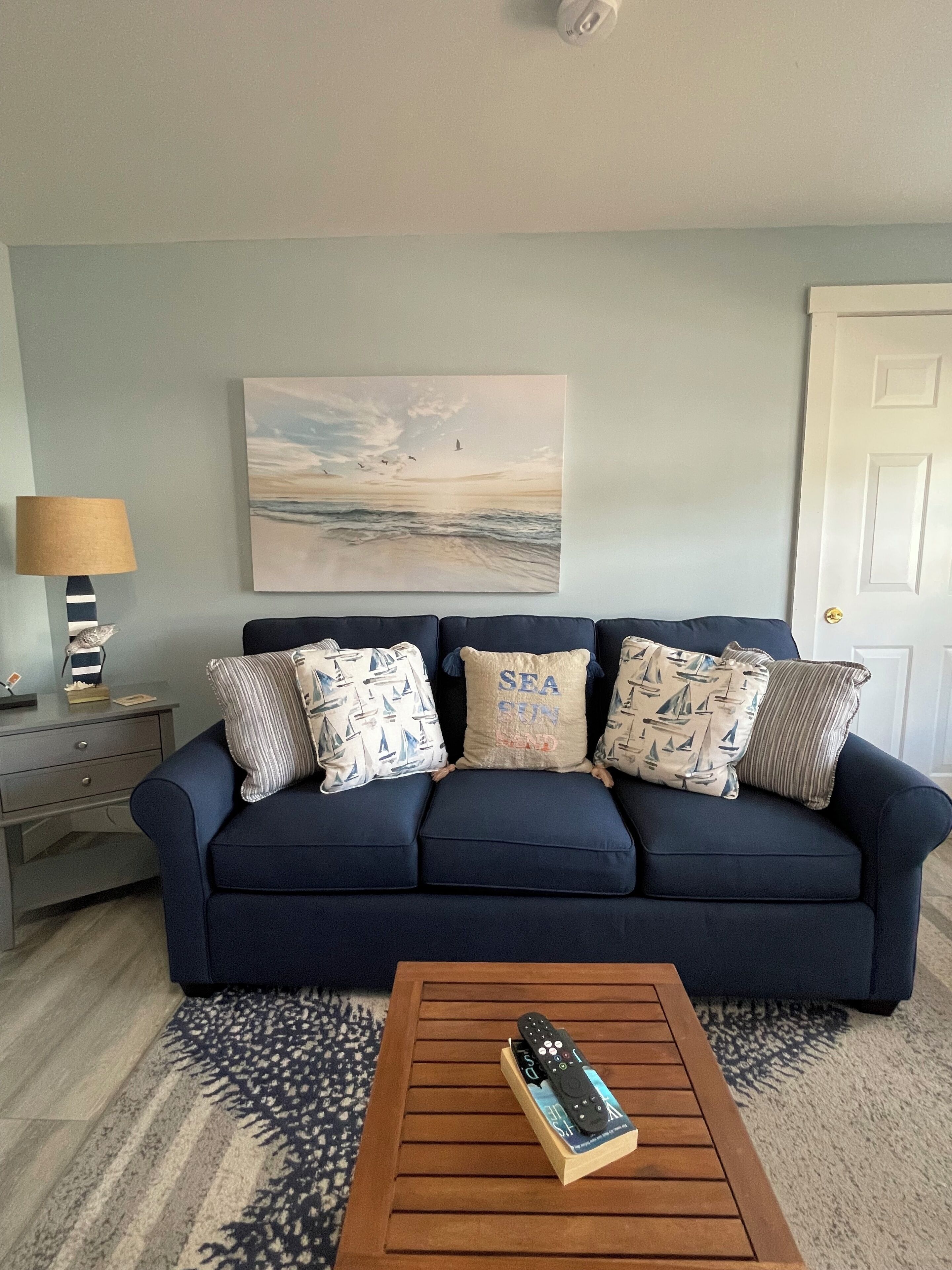 A living room with a navy blue sofa, patterned pillows, a painting of a seascape, a lamp, and a coffee table with a remote control.