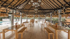 An open-air pavilion with wooden tables and benches, a thatched roof, and a view of a swimming pool. - Hacienda Oro Verde (San Jerónimo)