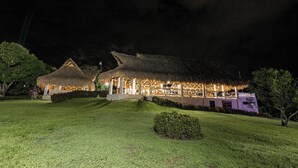 A thatched-roof building with a well-lit outdoor area, surrounded by greenery. - Hacienda Oro Verde (San Jerónimo)