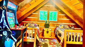 A cozy attic bedroom with wooden beams, a ceiling fan, two windows with green foliage outside, a small Christmas tree, and a retro arcade machine. - Hidden Treasure (Sevierville)
