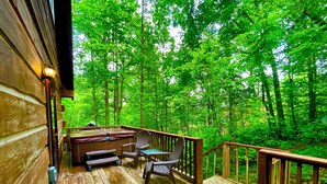 A wooden deck with outdoor seating, a hot tub, and a wooden railing overlooking a forest. - Hidden Treasure (Sevierville)