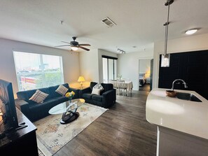 Een moderne woonkamer met een bank, salontafel en televisie. Er is een eethoek met een tafel en stoelen. De keuken heeft een spoelbak en een raam.