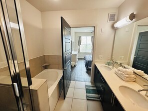 Een moderne badkamer met een bad, een wastafel en een spiegel. Een deur leidt naar een slaapkamer met een bed en een raam.