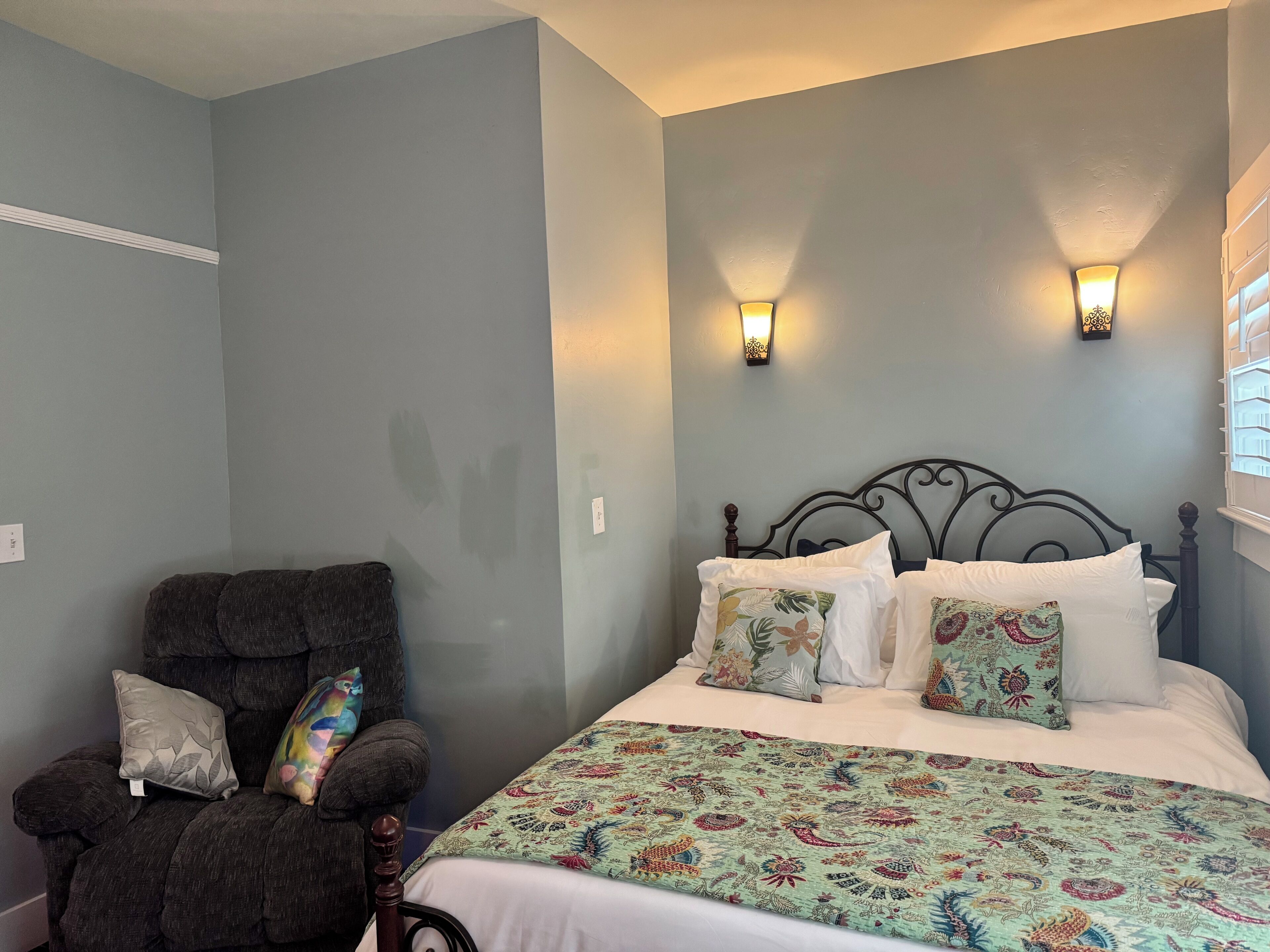 A bedroom with a bed, two wall sconces, a chair, and a patterned bedspread.