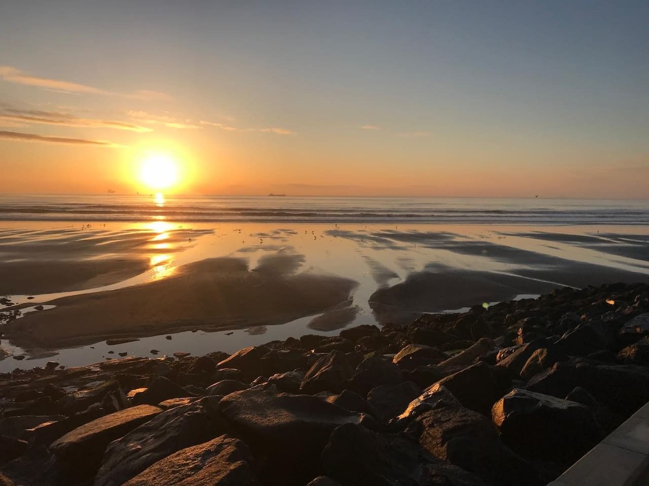 Eine Strandansicht mit Felsen und ruhigem Wasser, das den Sonnenuntergang spiegelt.